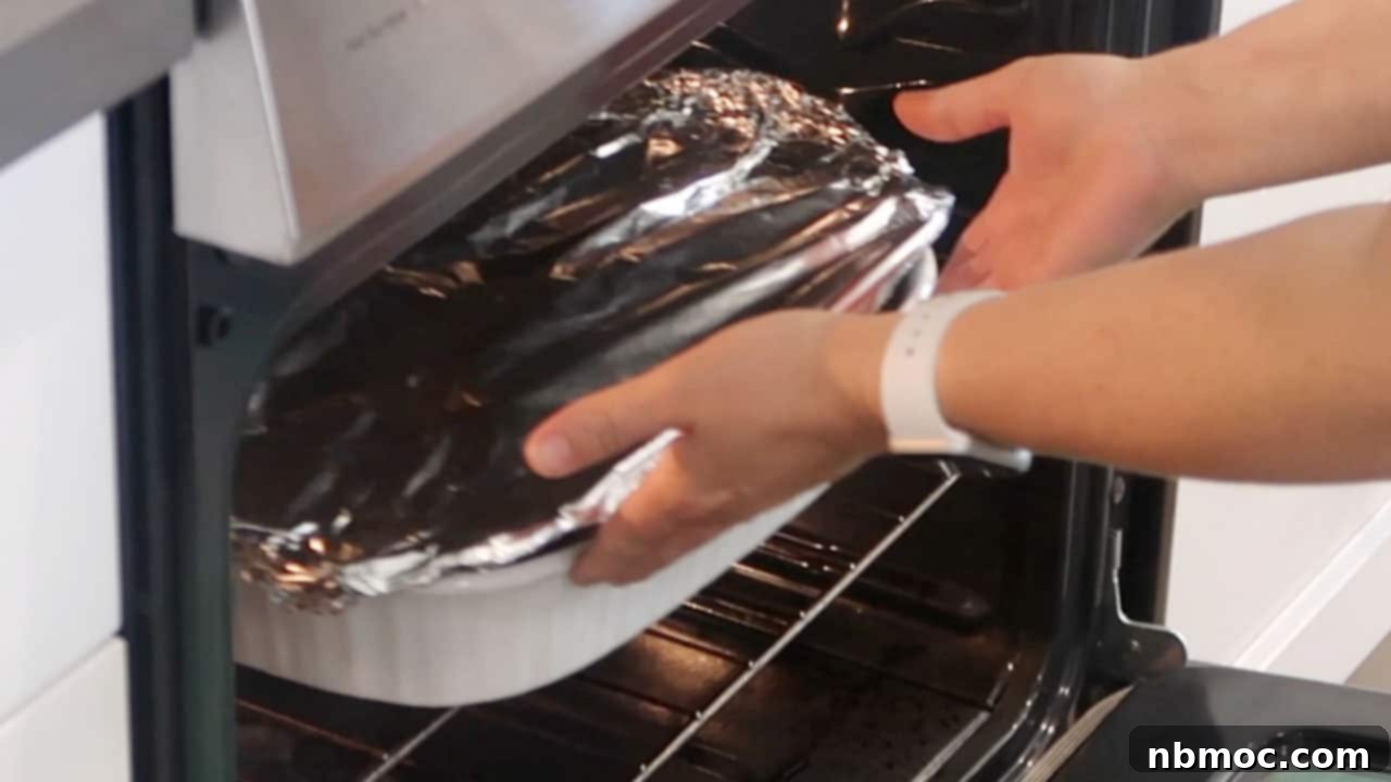Hands carefully placing a baking dish, covered with aluminum foil, into a preheated oven for roasting.