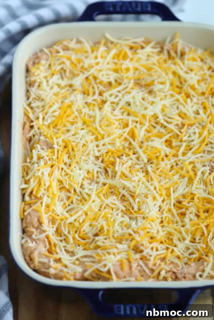 Uncooked cream cheese refried bean dip, topped with a generous layer of shredded cheese, in a blue baking dish before baking.