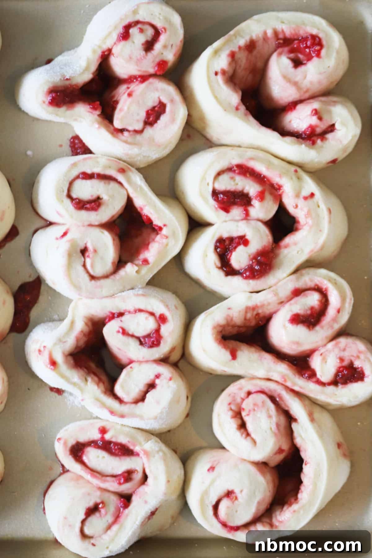 Raspberry sweet rolls prepared before baking, resting on a large baking sheet, ready for the oven to become golden brown and fluffy.
