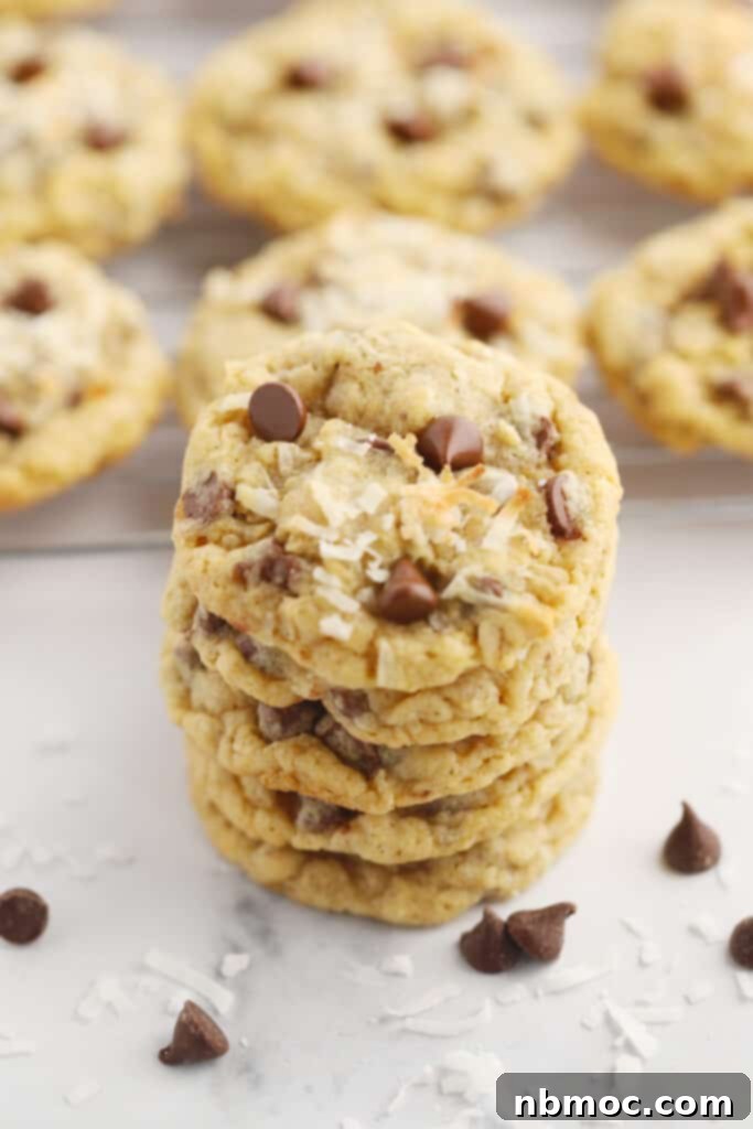 Simply Delicious Chewy Cowboy Cookies 2 Chocolate Chip cowboy cookies stacked on parchment paper with a few chocolate chips and flakes of coconut.