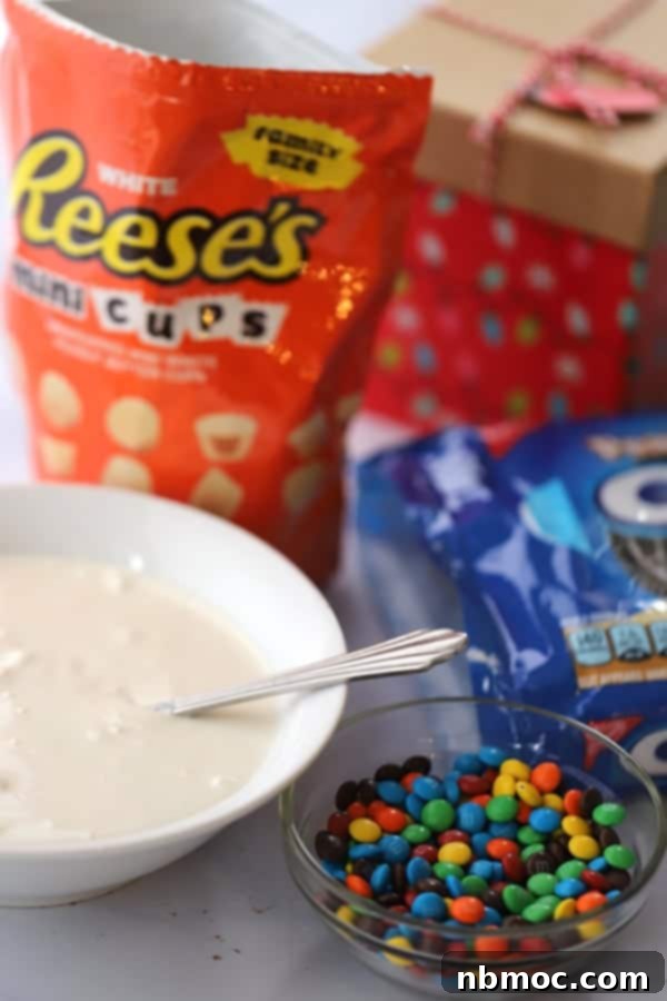 Ingredients for making festive chocolate covered oreos, including white chocolate melting wafers, colorful M&Ms, and mini Reese's peanut butter cups for ornament toppers.