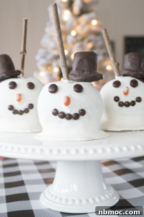Three Snowman Christmas Caramel Apples on a cake stand on a table with a checkered tablecloth, ready for a holiday party.