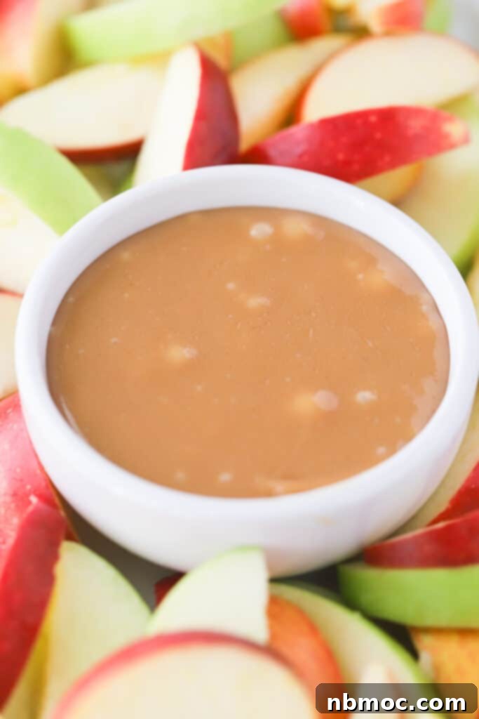 A bowl of glossy, golden caramel dipping sauce for apples, beautifully surrounded by an assortment of freshly sliced red and green apples, inviting a delicious treat.