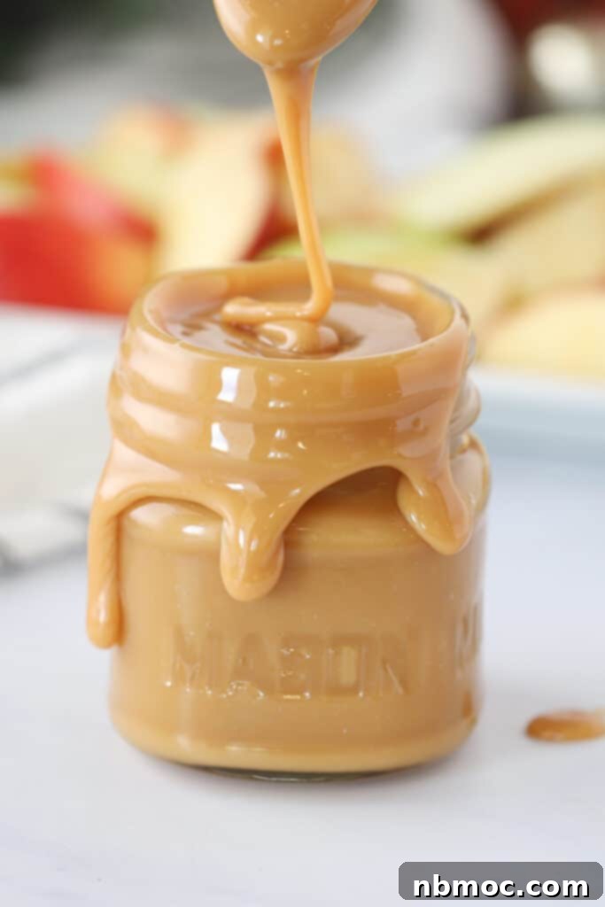 A small mason jar overflowing with a rich, golden, gooey caramel apple dipping sauce, beautifully presented.