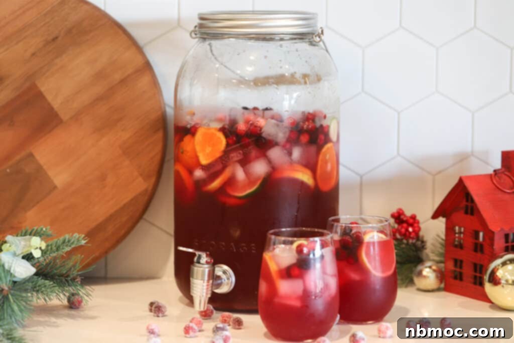 Festive Elixir 4 A table with a large drink dispenser and two individual glasses full of red punch, garnished with sliced fruit and cranberries.