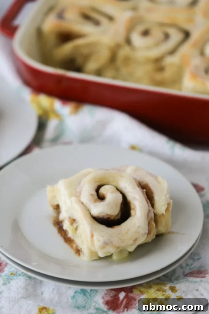 A perfectly baked, golden-brown homemade cinnamon roll generously covered with smooth cream cheese frosting, served on an elegant white serving plate, ready to be enjoyed.
