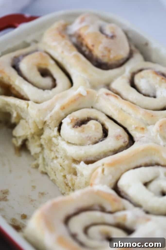 A close-up shot of a baked homemade cinnamon roll, generously covered with a thick layer of white cream cheese frosting, showcasing its inviting texture and warmth on a white plate.