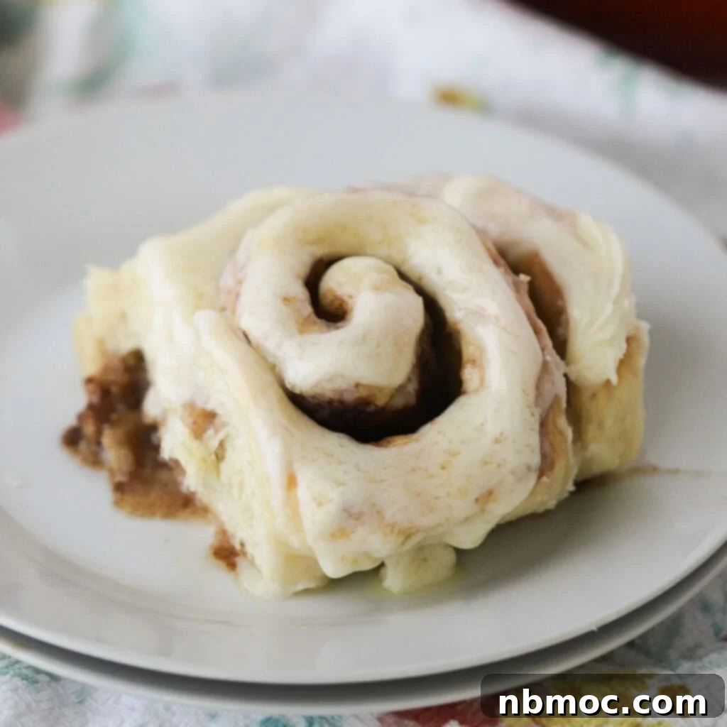 A single, perfectly baked cinnamon roll with a generous swirl of cinnamon filling and a thick layer of cream cheese frosting, presented on a simple white plate, highlighting its delicious homemade quality.