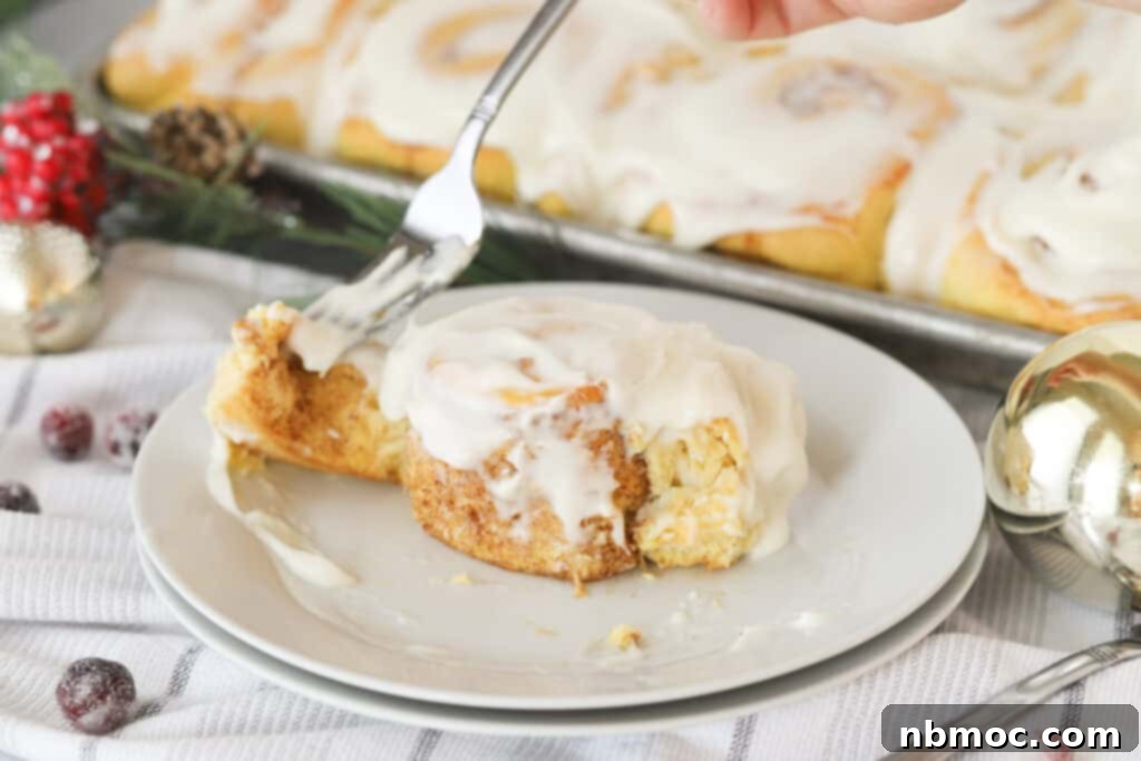 A close-up shot of a pumpkin cinnamon roll being eaten with a fork, with a full baking dish of frosted rolls blurred in the background, showcasing their deliciousness.