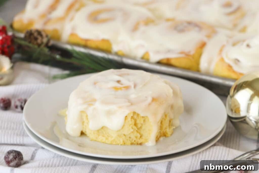 A close-up of a frosted Lemon Sweet Roll on a white plate, with a pan of more sweet rolls blurred in the background.