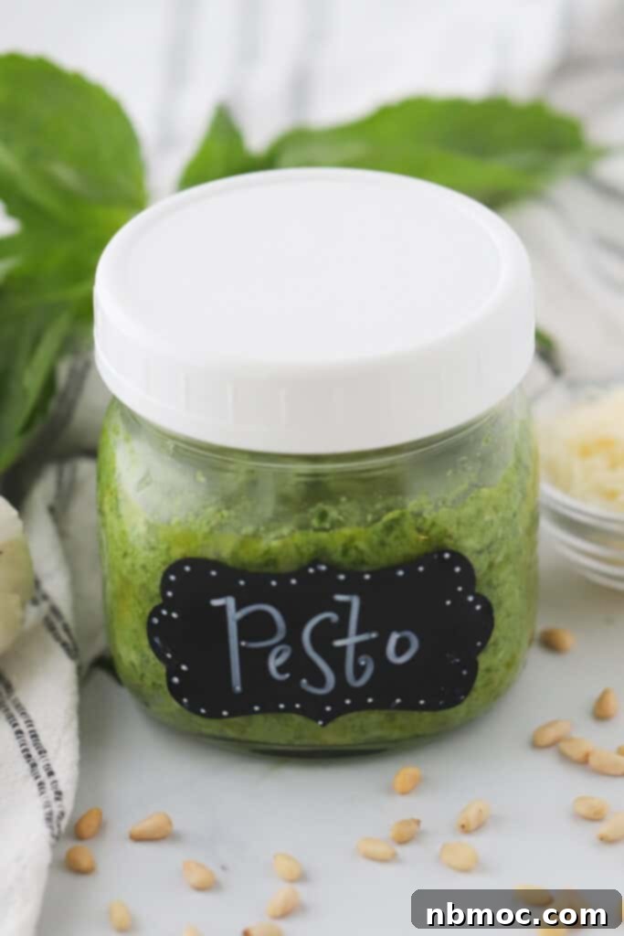 Homemade Pesto in a small jar on a table surrounded by pine nuts and basil. This recipe shows you how to make fresh pesto at home.