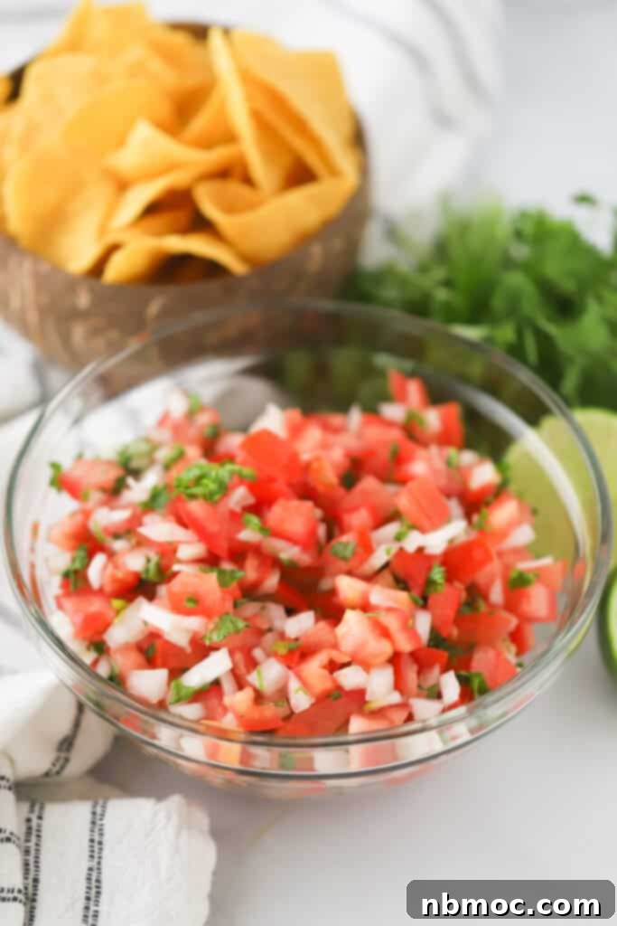 A glass serving bowl full of our simple Pico de Gallo recipe, with chips, fresh limes and cilantro in the background.