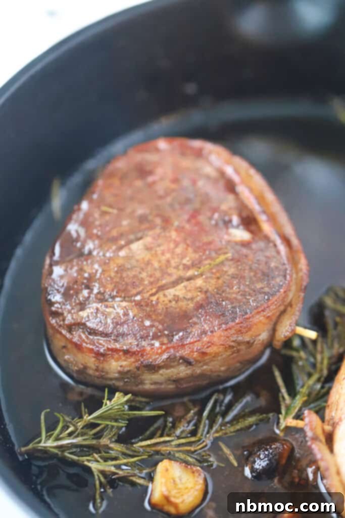 Perfectly cooked Bacon Wrapped Filet Mignon sizzling in a cast iron skillet with fresh rosemary and garlic cloves. This image highlights how to cook bacon wrapped filet, steak bacon wrapped, and filet mignon wrapped in bacon for an exquisite meal.