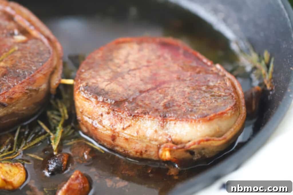A perfectly pan-seared bacon wrapped filet mignon resting in a cast iron skillet alongside aromatic rosemary and garlic, ready to be served. This image showcases delicious bacon wrapped filet mignons, filet mignon cut, bacon wrapped steaks, and how to cook filet mignon wrapped in bacon.