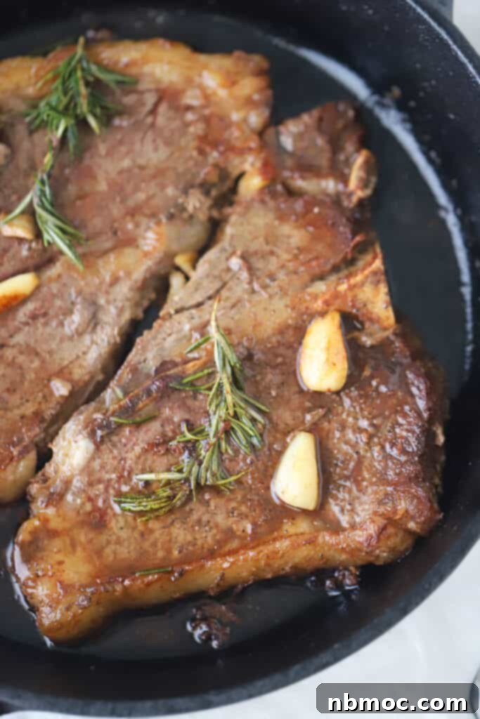 Pan seared steak in a cast iron skillet topped with rosemary and garlic cloves, ready for the oven finish.