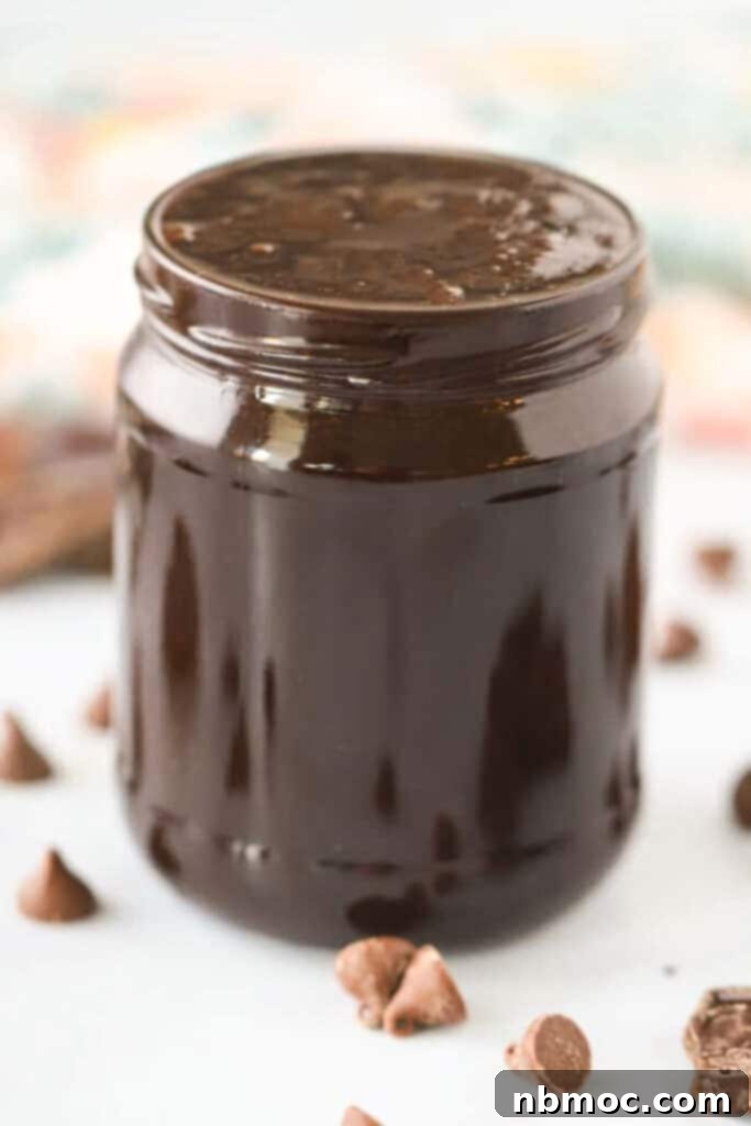 A glass jar full of chocolate sauce recipe on a table surrounded by mini chocolate chips and a spoon.