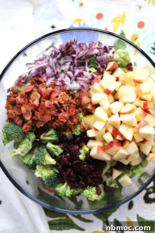 A refreshing glass bowl of classic Broccoli Salad with Bacon, featuring raw broccoli florets, crispy bacon, red onions, diced apples, and dried cranberries.