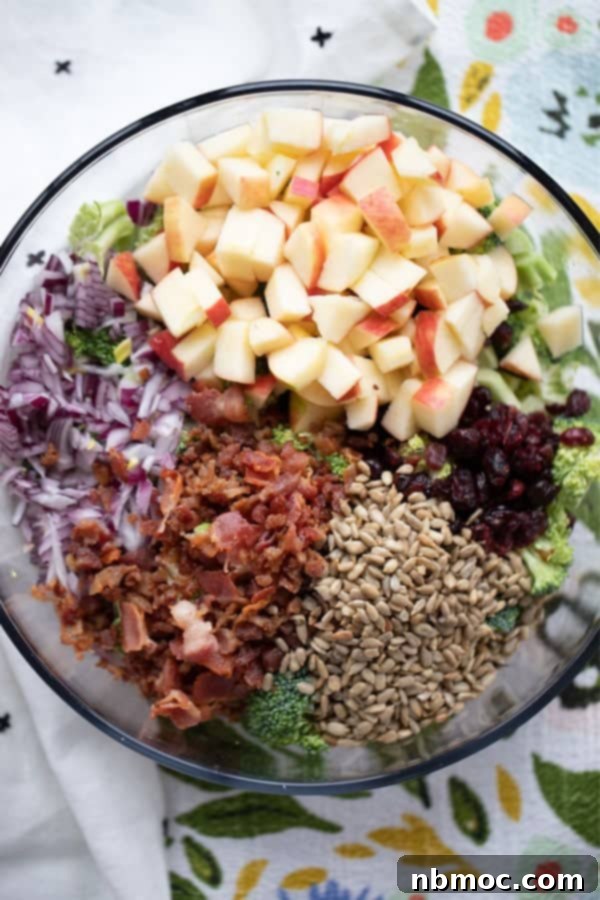 A large mixing bowl filled with raw broccoli, crispy bacon bits, diced apples, red onion, dried cranberries, and sunflower seeds, ready for the dressing.