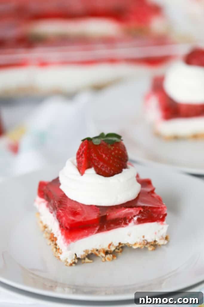 A beautifully presented slice of strawberry pretzel salad on a white serving plate, elegantly topped with a dollop of whipped cream and garnished with fresh, vibrant berries.