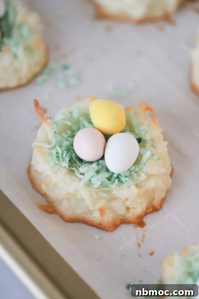 Adorable Coconut Macaroon Easter Nests 2 Coconut Birds Nest cookies topped with green colored shredded coconut and candy eggs on a baking sheet.