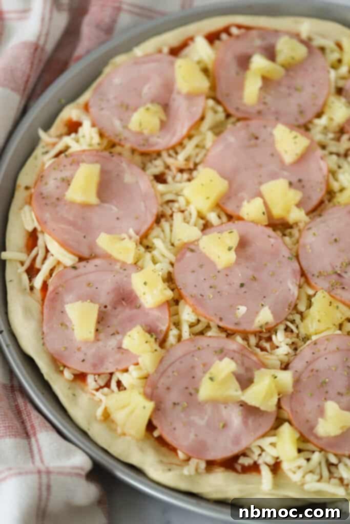 Best Hawaiian Pizza Recipe 5 Pizza dough on a pizza pan topped with Canadian bacon, shredded cheese, fresh pineapple and seasoning.