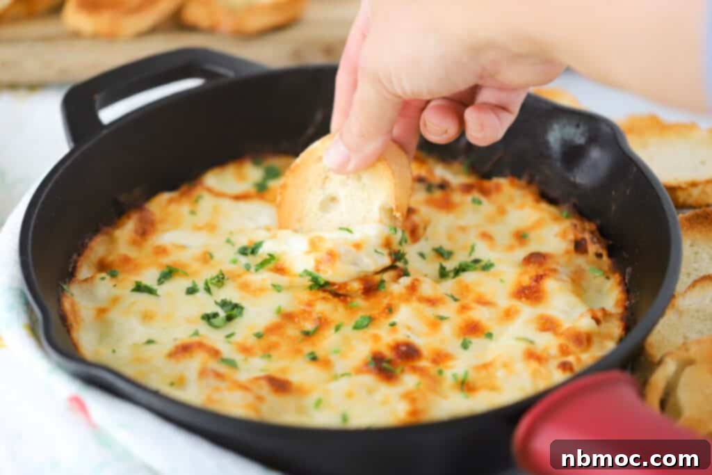 A cast iron skillet full of baked Alfredo dip, with a hand dipping a slice of bread into it.