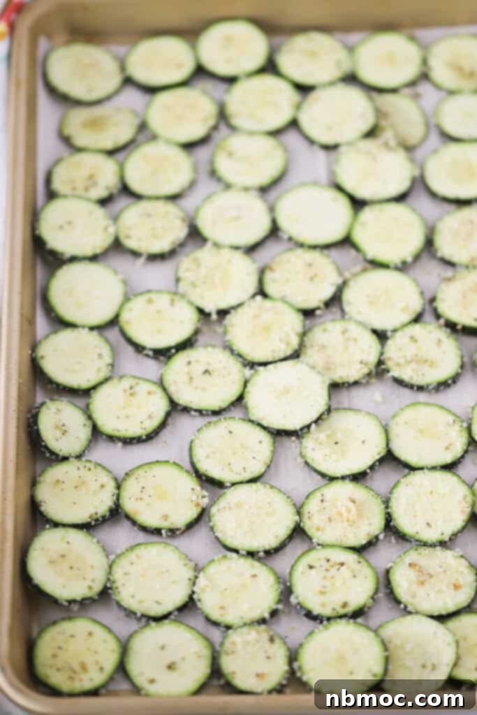 A sheet tray lined with parchment paper and a single layer of thinly sliced zucchini, topped with seasoning and Parmesan cheese for making baked zucchini chips.