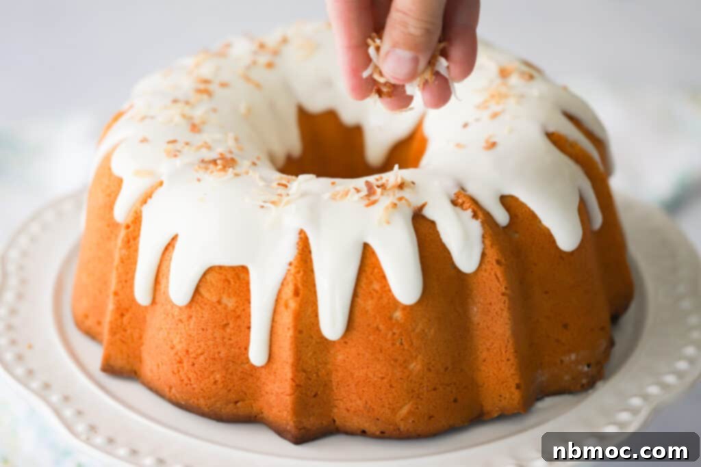 A hand sprinkling toasted coconut over a glazed bundt cake on a white serving plate. cream cheese pound cake with coconut. 