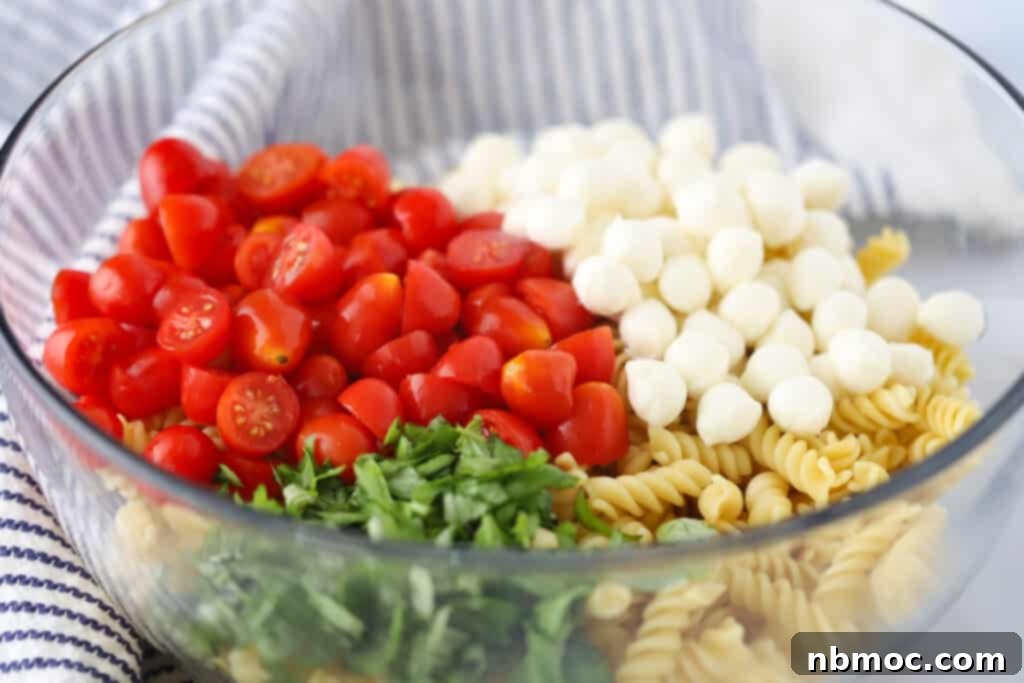 A glass mixing bowl filled with ingredients to make this mozzarella ball pasta salad, including cooked pasta, mozzarella balls, cherry tomatoes and fresh basil.