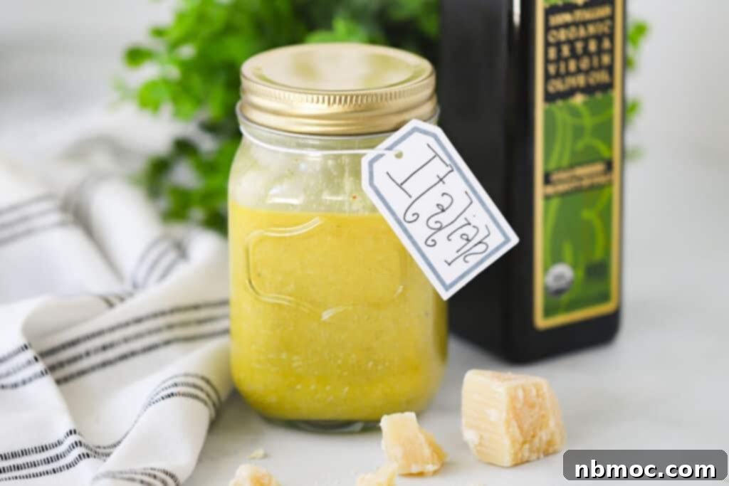 Zesty Italian Vinaigrette 4 A mason jar filled with creamy homemade Italian dressing, adorned with a "Italian" tag, rests on a rustic table beside a bottle of olive oil, a wedge of fresh Parmesan cheese, and a kitchen towel, highlighting the ease of making Italian Dressing at home.