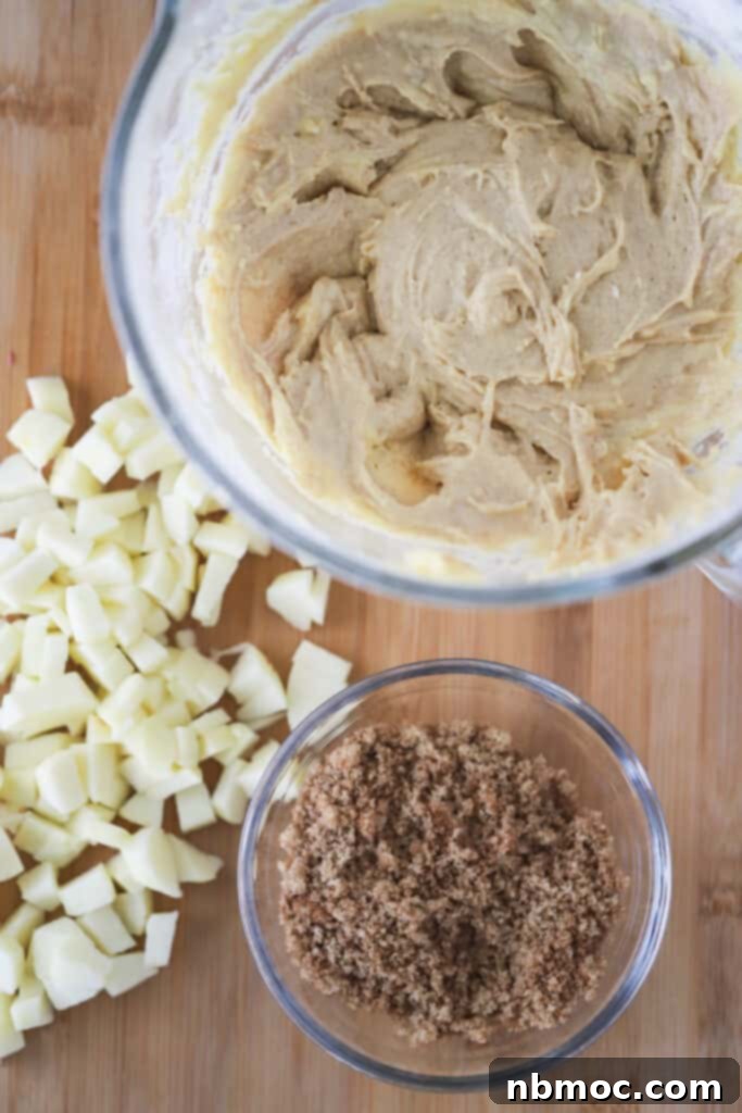 A tabletop scene showing a bowl of batter, a cup of cinnamon sugar, and a pile of chopped apples, all prepared for making this delicious apple cinnamon bread recipe.