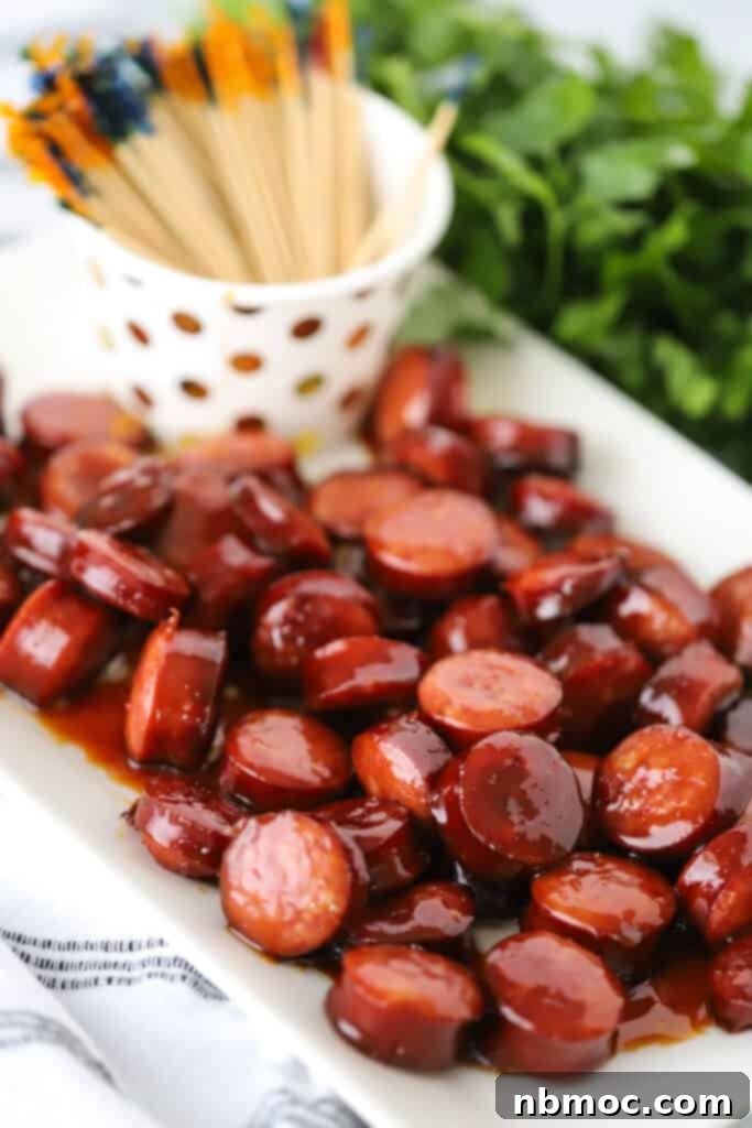 A beautifully arranged white serving platter filled with glistening sweet kielbasa, accompanied by a cup of decorative toothpicks.