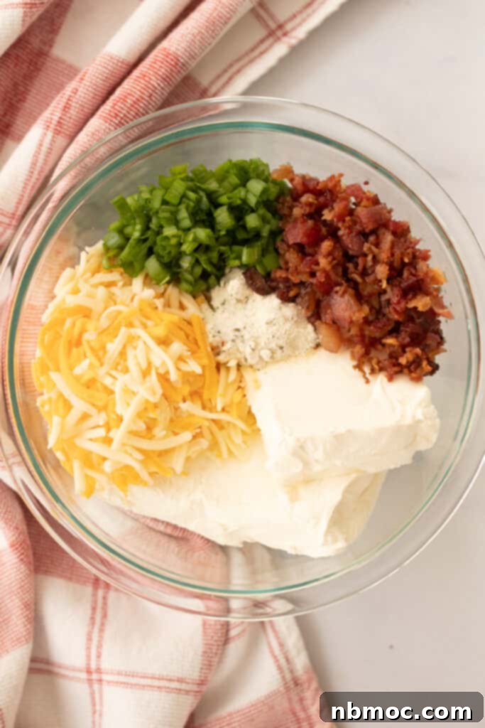 A glass mixing bowl filled with fresh ingredients for a Hidden Valley Ranch Cheese Ball, including cream cheese, cheddar, bacon, and green onions.