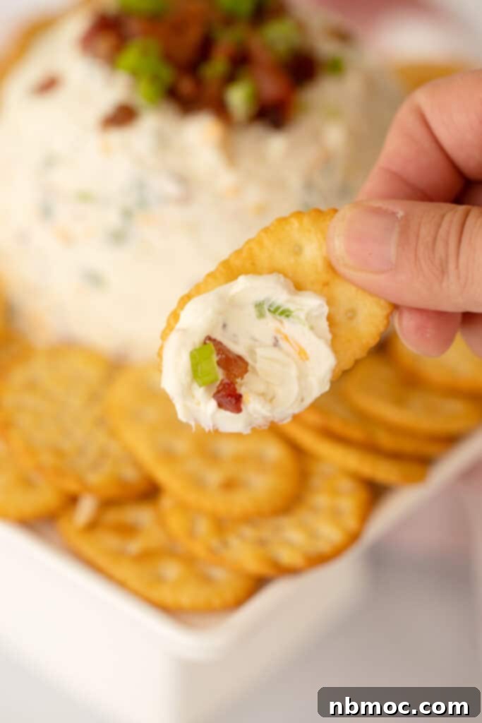 A hand dipping a crispy cracker into a rich and creamy Bacon Ranch Cheese Ball, highlighting its inviting texture.