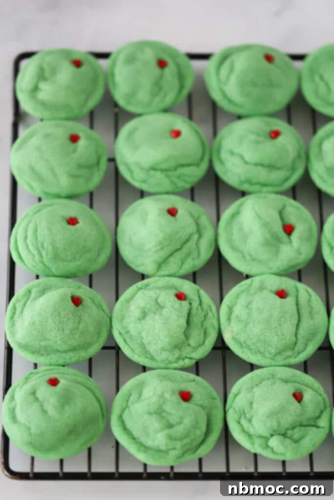 Green Mean Christmas Treats 2 A wire rack full of vibrant green Grinch Sugar Cookies, each featuring a small, iconic red candy heart, ready for festive enjoyment.