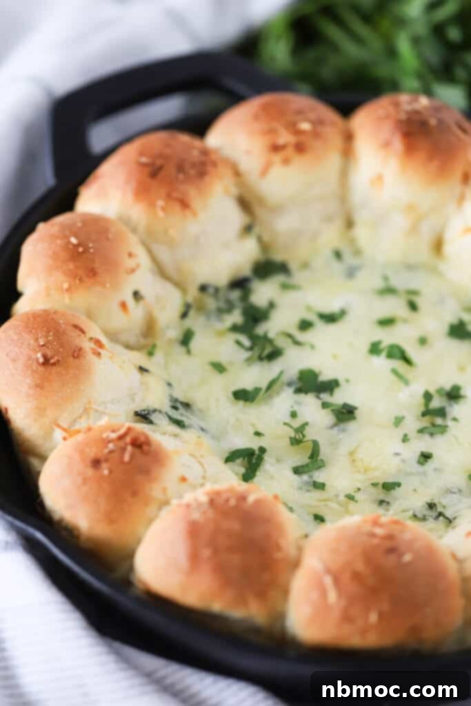 A skillet with a ring of perfectly baked, golden dinner rolls surrounding a bubbling, cheesy spinach dip in the center.