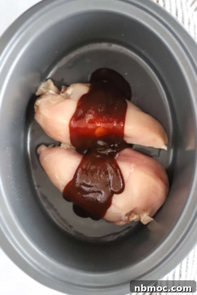 Raw chicken breasts nestled in a slow cooker, generously covered with BBQ sauce and chicken broth, ready for slow cooking. This image demonstrates the initial step for making easy slow cooker BBQ chicken sandwiches.