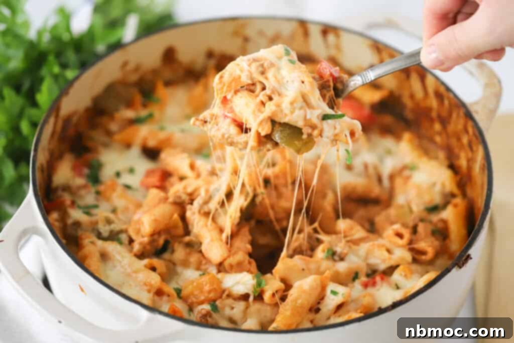 A Dutch oven full of this sausage and pepper pasta bake with a hand holding a spoon that is taking out a large scoop.