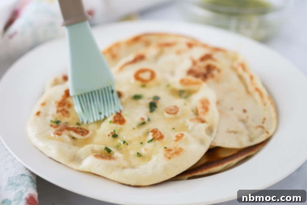 A freshly cooked naan bread on a white plate, being brushed with melted garlic butter for an irresistible finish. Best homemade naan bread recipe.