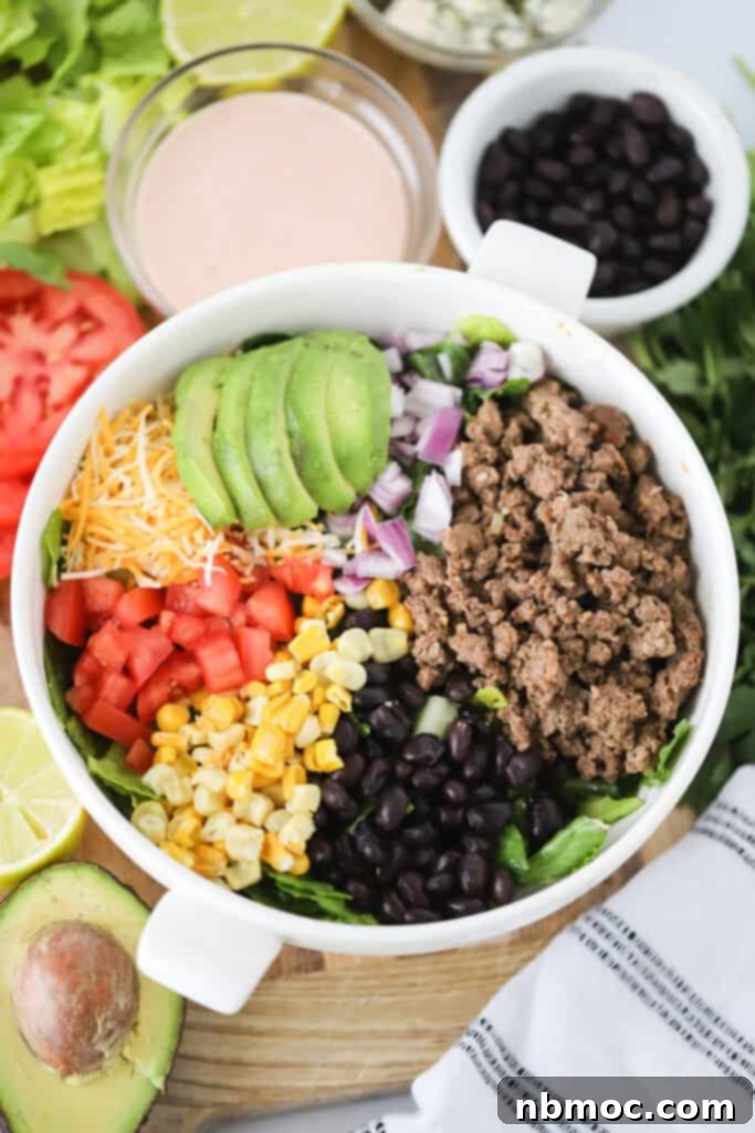 A serving bowl full of easy taco salad ingredients like ground beef, corn, avocadoes, and beans with a side of southwest ranch dressing.