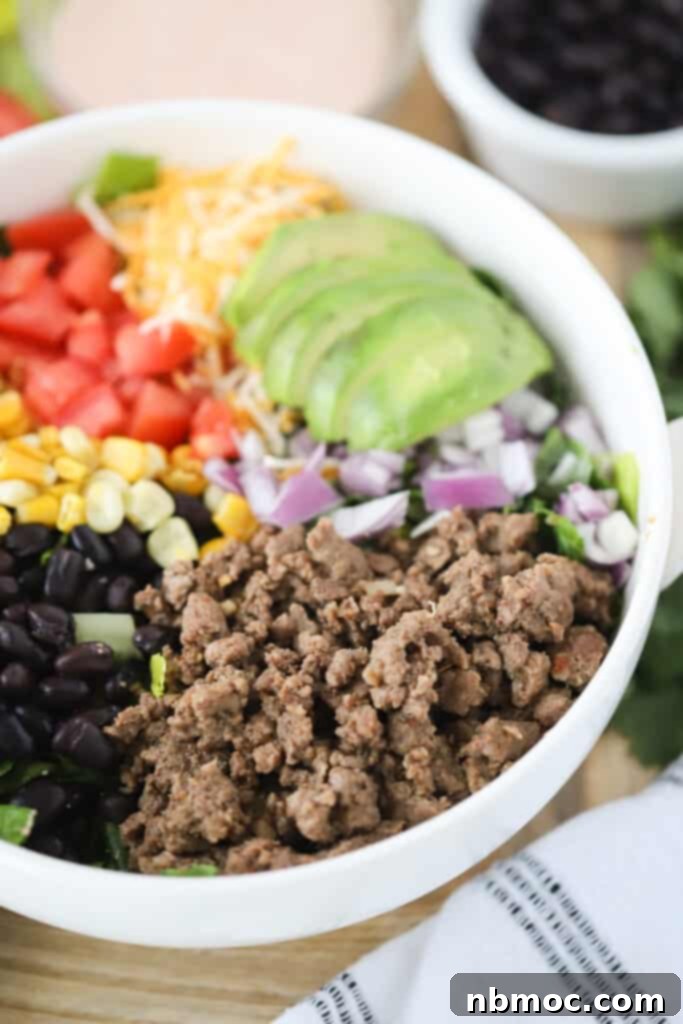 A bowl full of ingredients to make easy Taco Salad including ground beef, sliced avocado, black beans, corn, tomatoes, and shredded cheese.