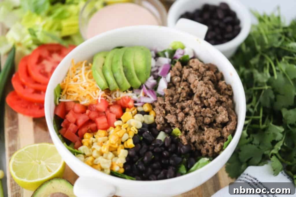 A bowl full of ingredients to make a salad including ground beef, black beans, corn, tomatoes, shredded cheese, and avocados.