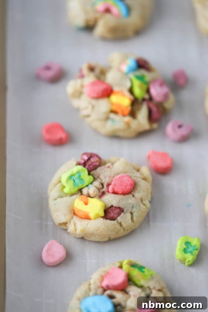 A plate of freshly baked Lucky Charms Cookies, topped with extra marshmallows, alongside a glass of milk and additional cookie ingredients. A perfect example of how to make a lucky charm cookie.