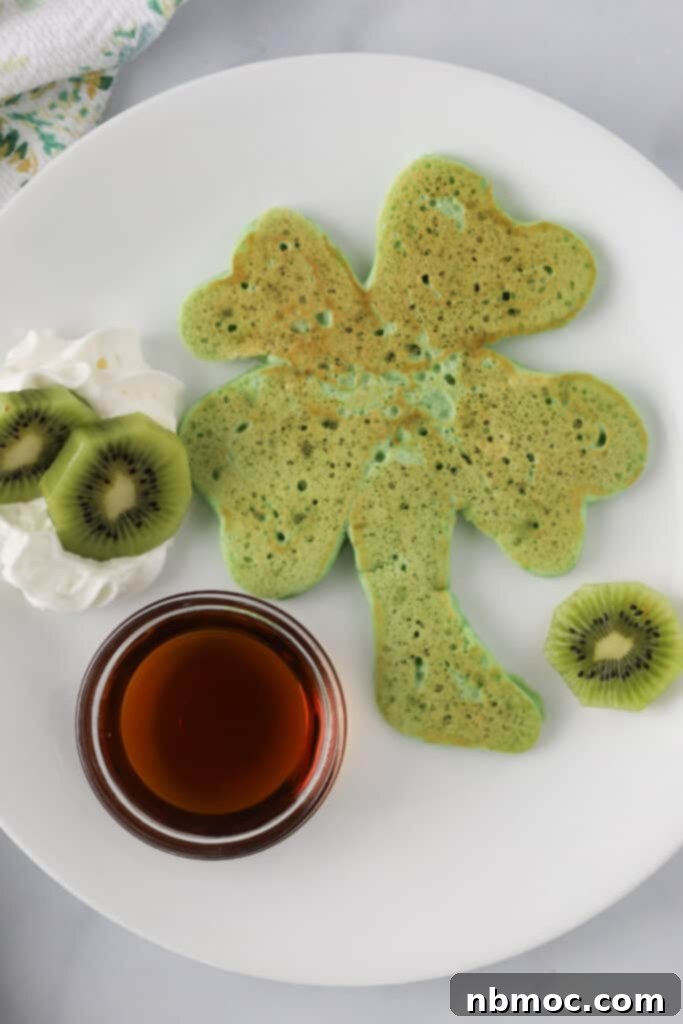 A white plate with a stack of vibrant green shamrock shaped St. Patrick's Day pancakes, accompanied by a generous drizzle of syrup and a side of fresh sliced kiwi. This festive breakfast is perfect for St. Patrick's Day.