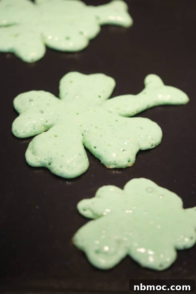 Vibrant green shamrock shaped pancakes gently cooking on a warm griddle, creating a festive atmosphere for St. Patrick's Day breakfast. These are perfect shamrock pancakes and a delightful green pancake recipe.