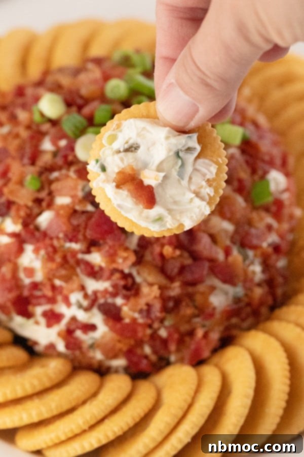 Kickin' Jalapeño Cheese Ball 2 A hand holding a cracker dipped into a creamy Jalapeno Popper Cheese Ball, with the full appetizer and additional crackers in the background, ready for serving.