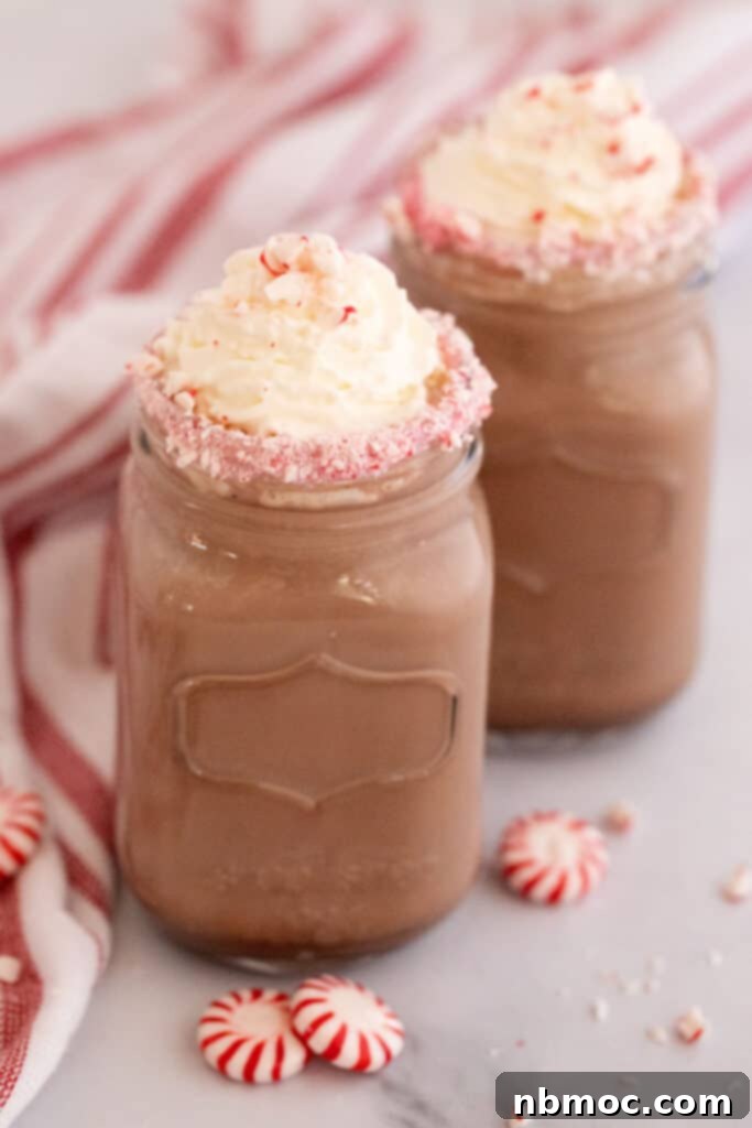 Mason jars filled with frothy crockpot hot chocolate, adorned with a festive peppermint candy rim and topped with whipped cream, ready for serving.