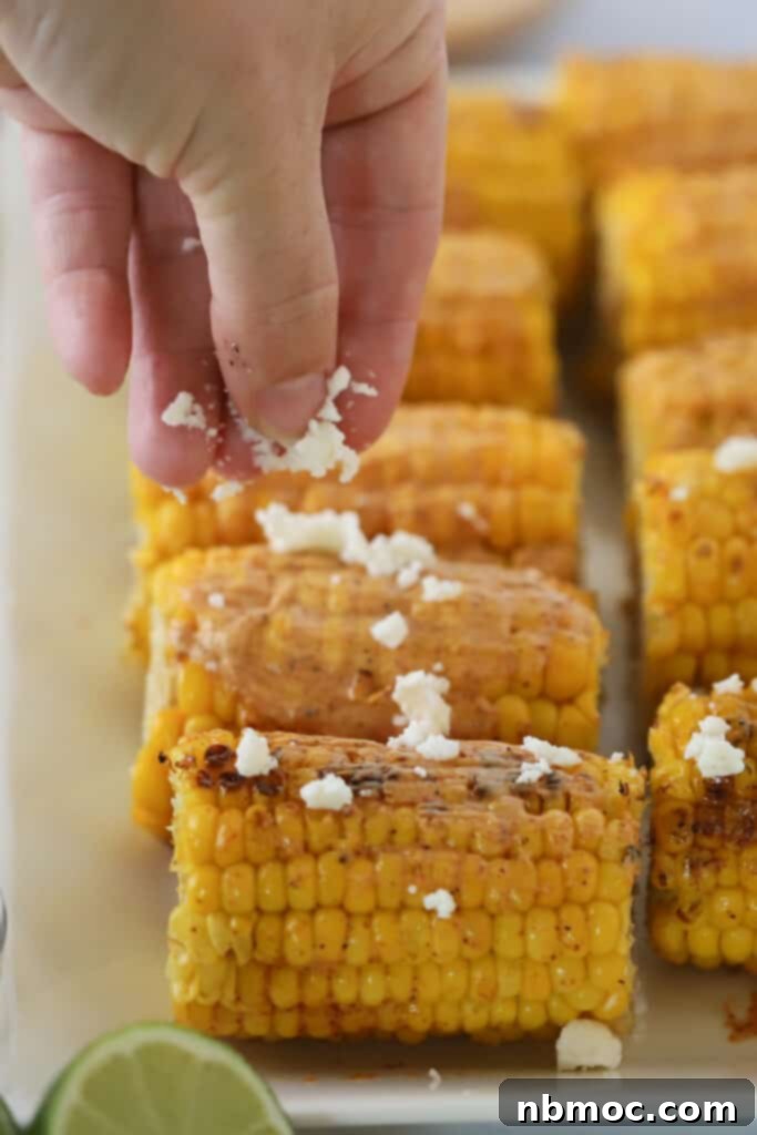 A hand sprinkling cotija cheese over the top of grilled corn on the cob.