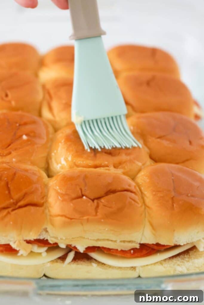 Close-up of a hand brushing melted butter and seasoning on the tops of Hawaiian rolls for pizza sliders before baking.