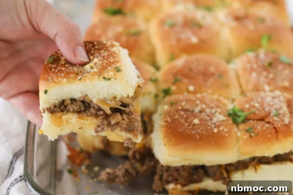 A hand reaching for a cheeseburger slider on a Hawaiian roll from a baking dish, highlighting their grab-and-go appeal.